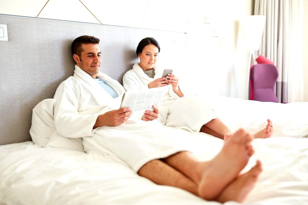 The image shows a couple relaxing in their bed while reading a newspaper and checking their mobile phone.
